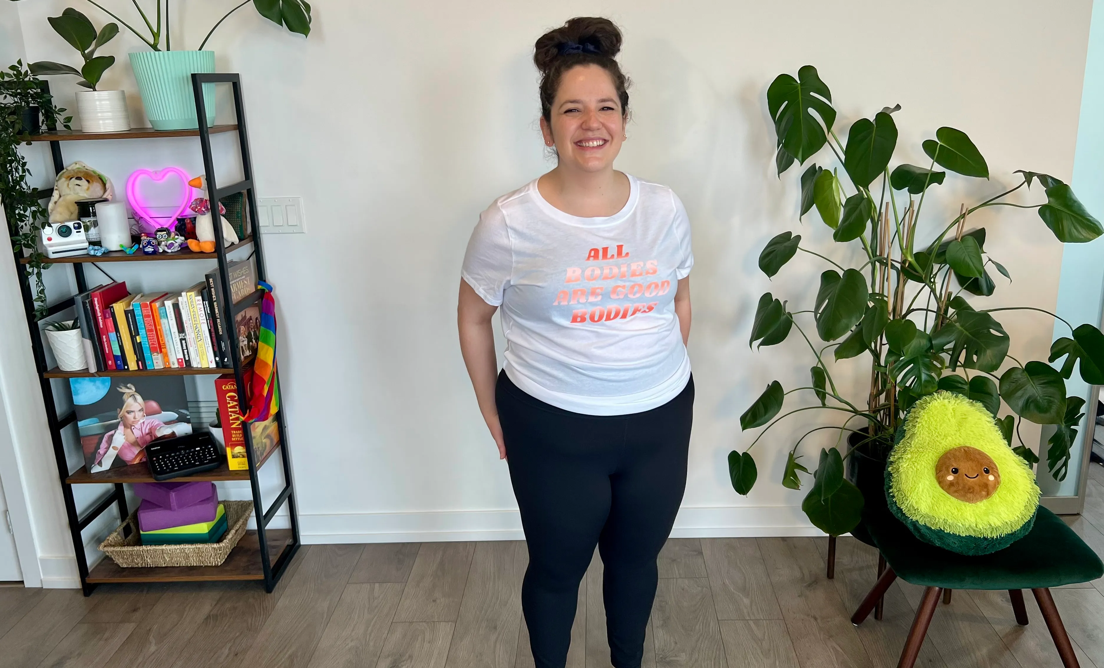 Rebecca Yaffa smiling in an All Bodies Are Good Bodies t-shirt in her bright, plant-filled home studio.